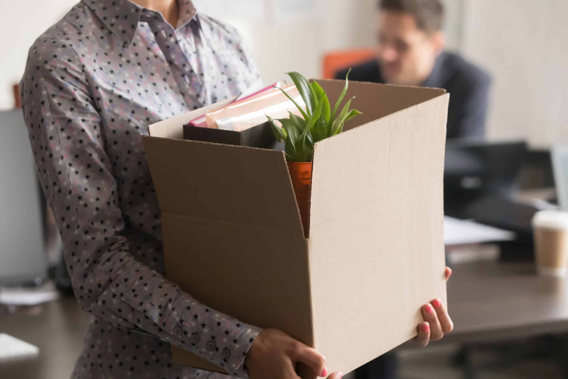 A person carries a box filled with office belongings, including a plant.