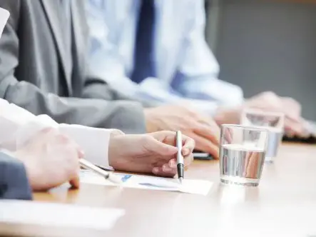 Business meeting. Attendees in suits sit around a table reviewing paperwork, and glasses of water are placed on the table.